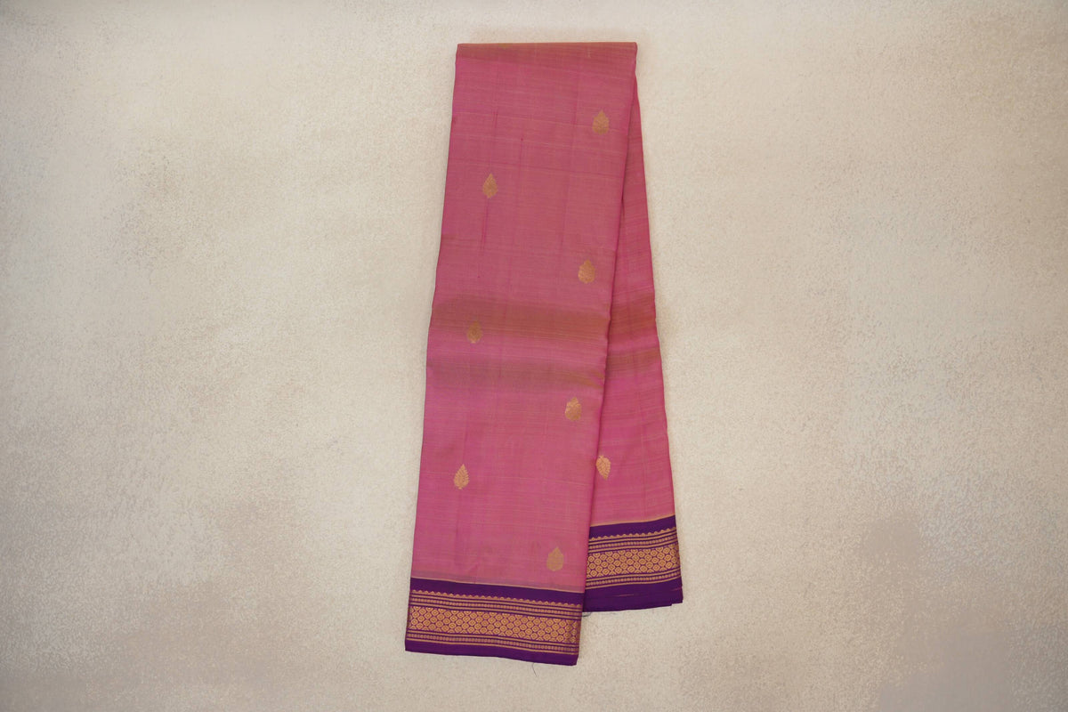 Folded rose pink Kanchipuram silk saree with scattered gold leaf-shaped motifs and a purple border woven with gold geometric patterns.