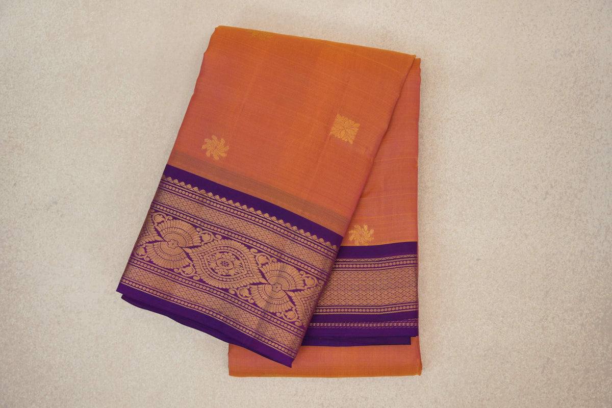 Folded light weight Kanchipuram silk saree with a warm pink and yellow body, small gold floral motifs and an ornate deep purple border.