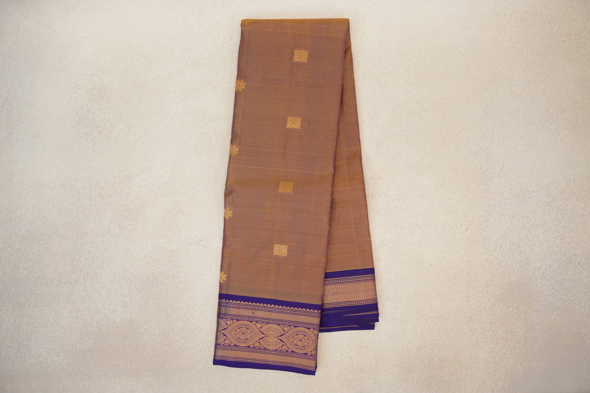 Folded Kanchipuram silk saree with a muted magenta and yellow body, small gold motifs, and violet purple borders with ornate gold detailing.