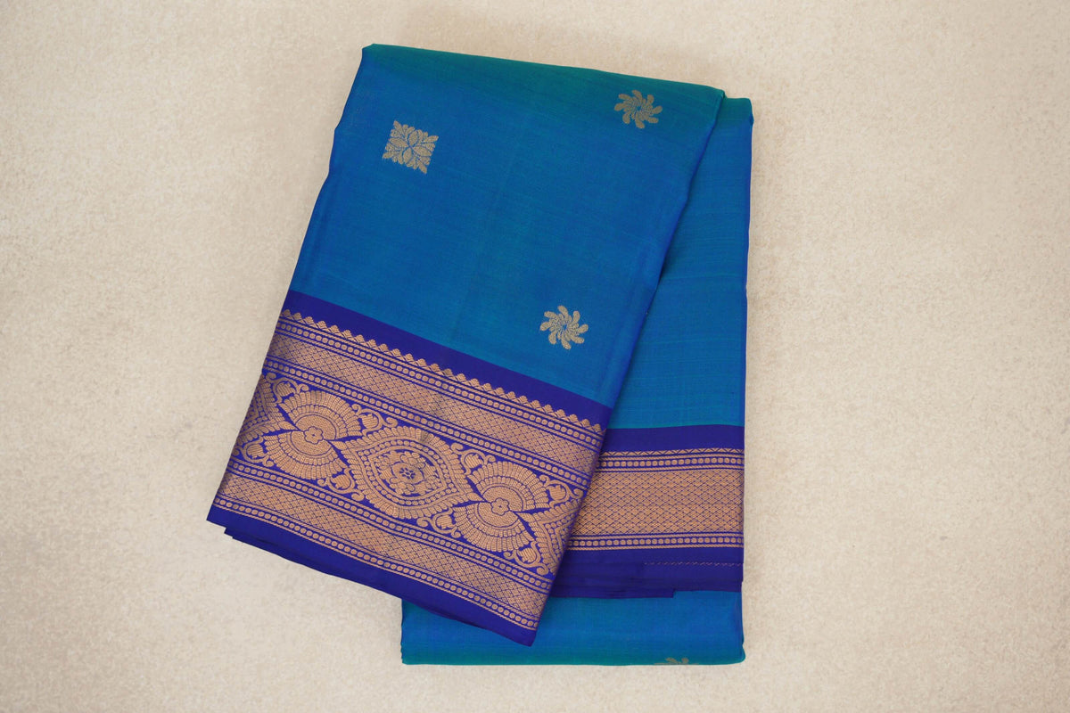Folded peacock blue lightweight Kanchipuram silk saree with small gold floral motifs and an ornate gold-patterned deep royal blue border.