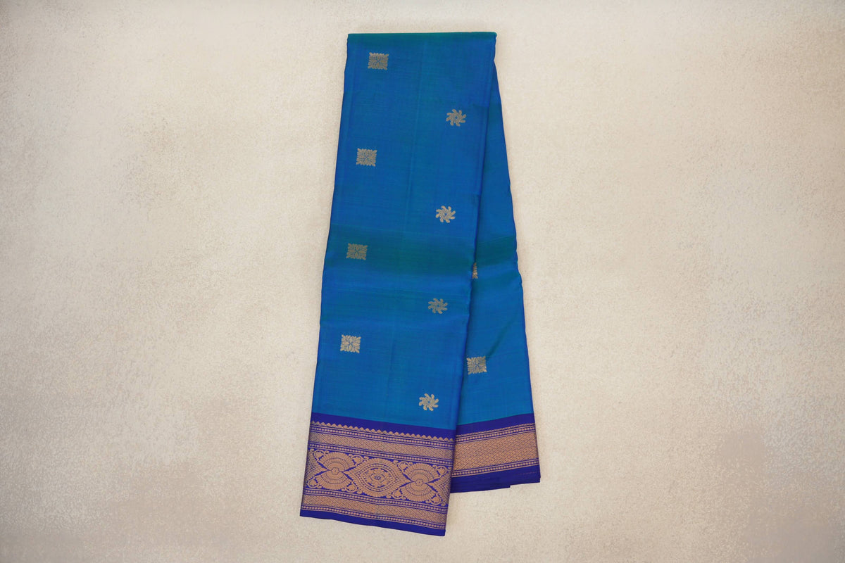 Folded peacock blue lightweight Kanchipuram silk saree with scattered gold motifs and a deep royal blue border with gold floral patterns.