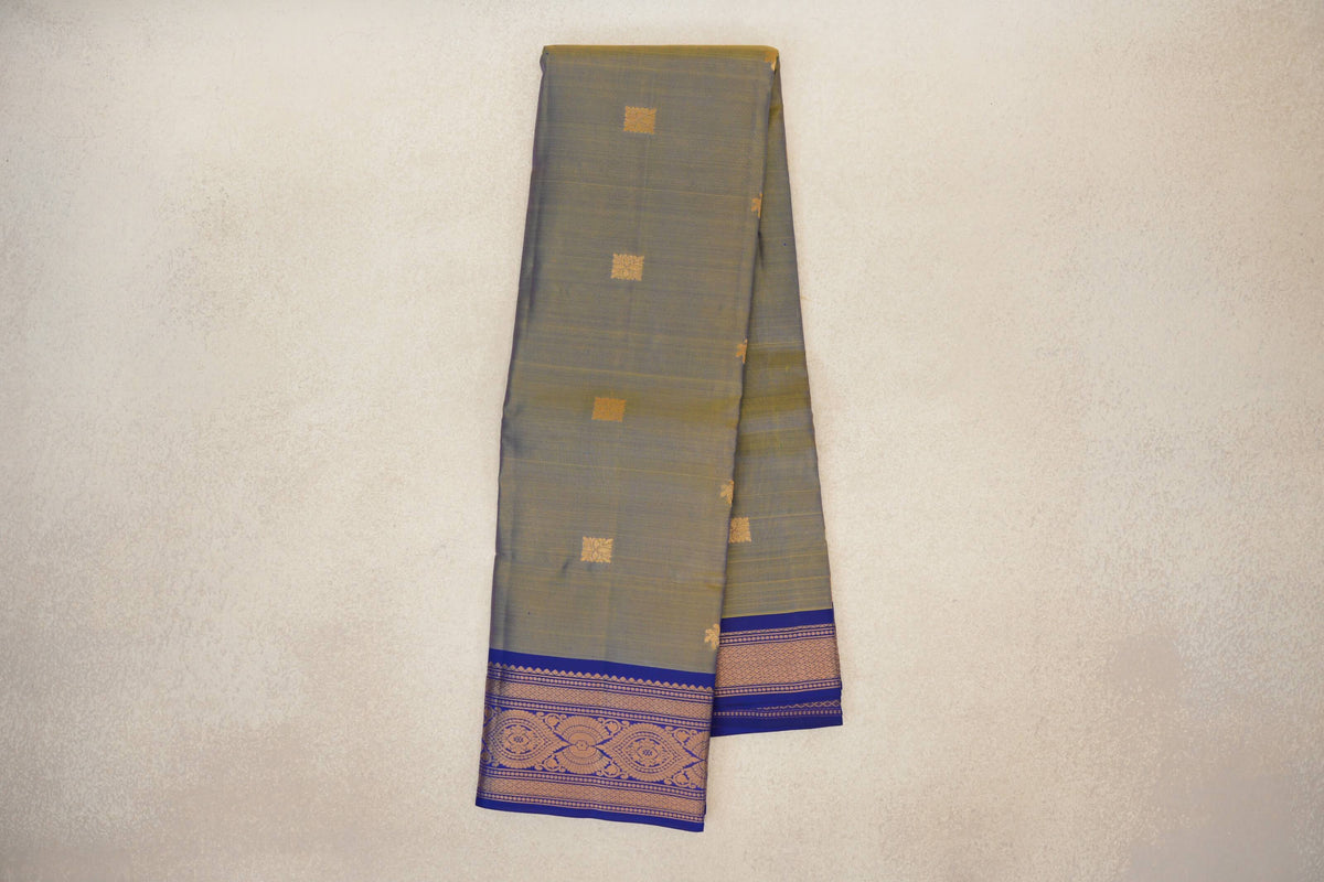 Folded light weight Kanchipuram silk saree with a muted yellow-violet body, small gold square motifs and patterned deep royal blue borders.