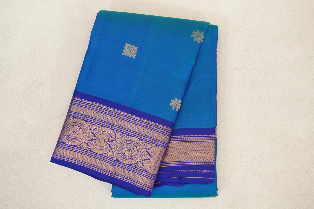 Folded peacock blue lightweight Kanchipuram silk saree with small gold floral motifs and an ornate deep royal blue border.