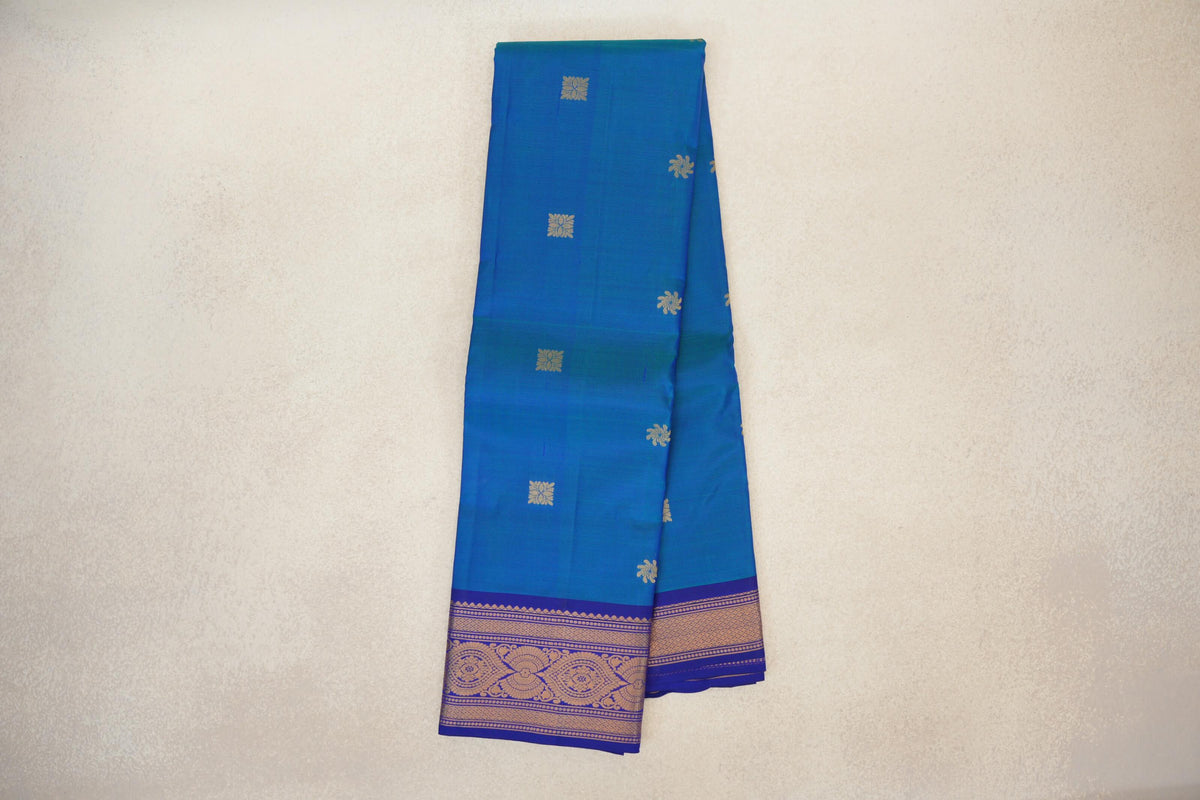 Folded peacock blue lightweight Kanchipuram silk saree with scattered gold floral motifs and a deep royal blue border with gold patterns.