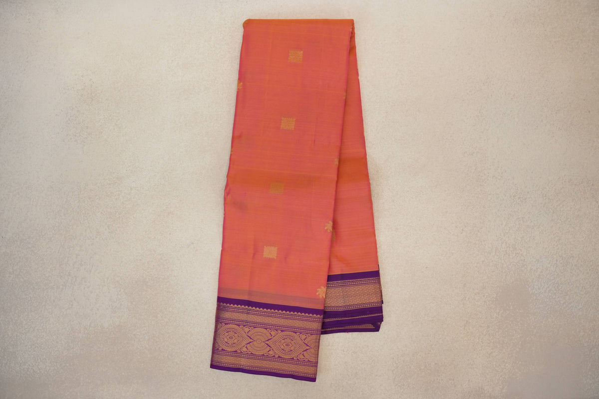 Folded Kanchipuram silk saree with a pink body, small gold square motifs and an ornate gold-patterned violet purple border.