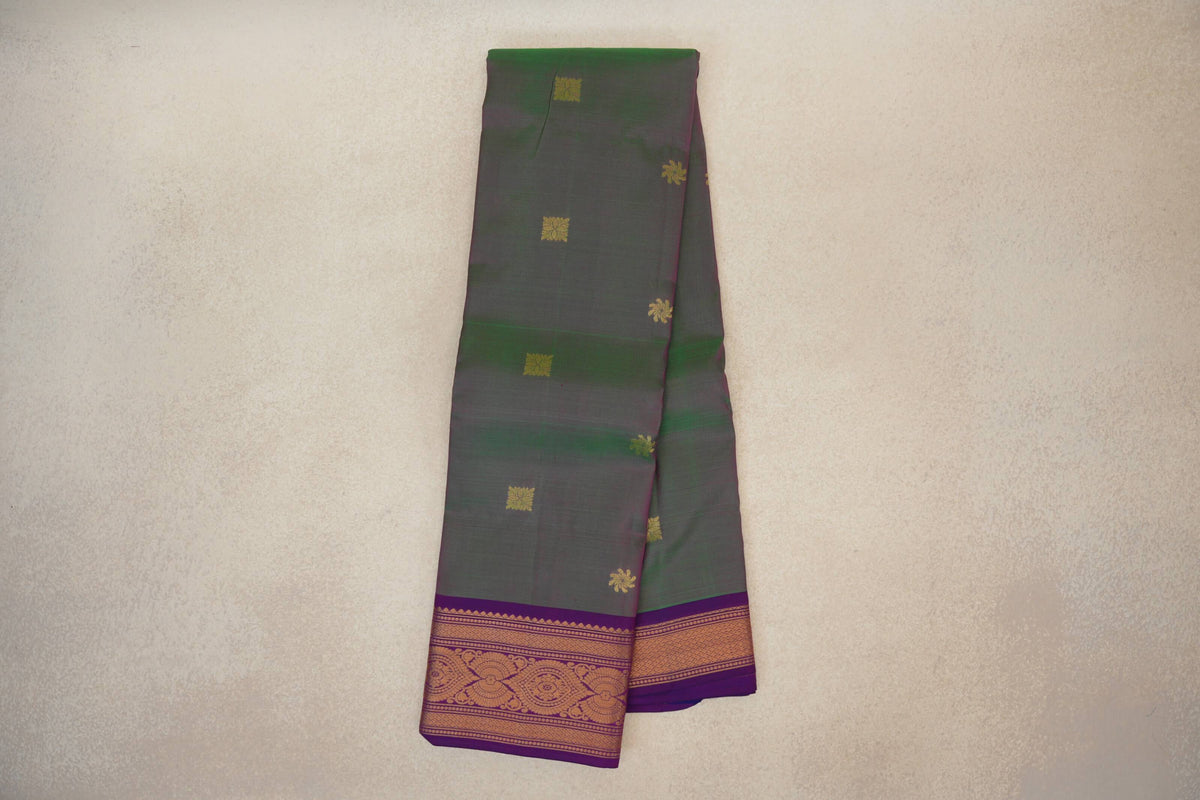 Folded light weight Kanchipuram silk saree with a muted green and pink body, scattered gold motifs and a patterned violet purple border.