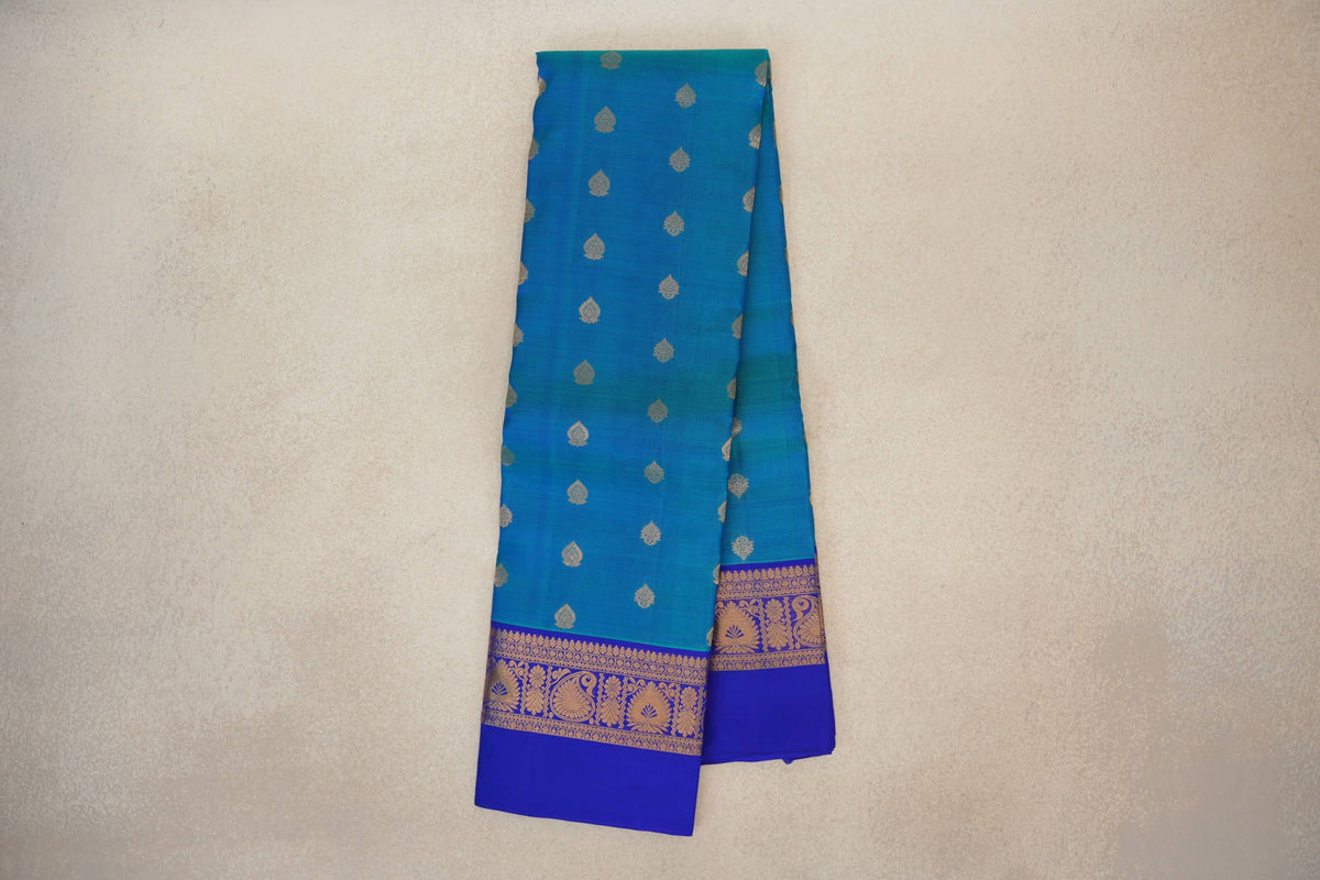 Folded peacock blue Kanchipuram silk saree with small gold-toned motifs and royal blue borders patterned with paisleys and flowers.