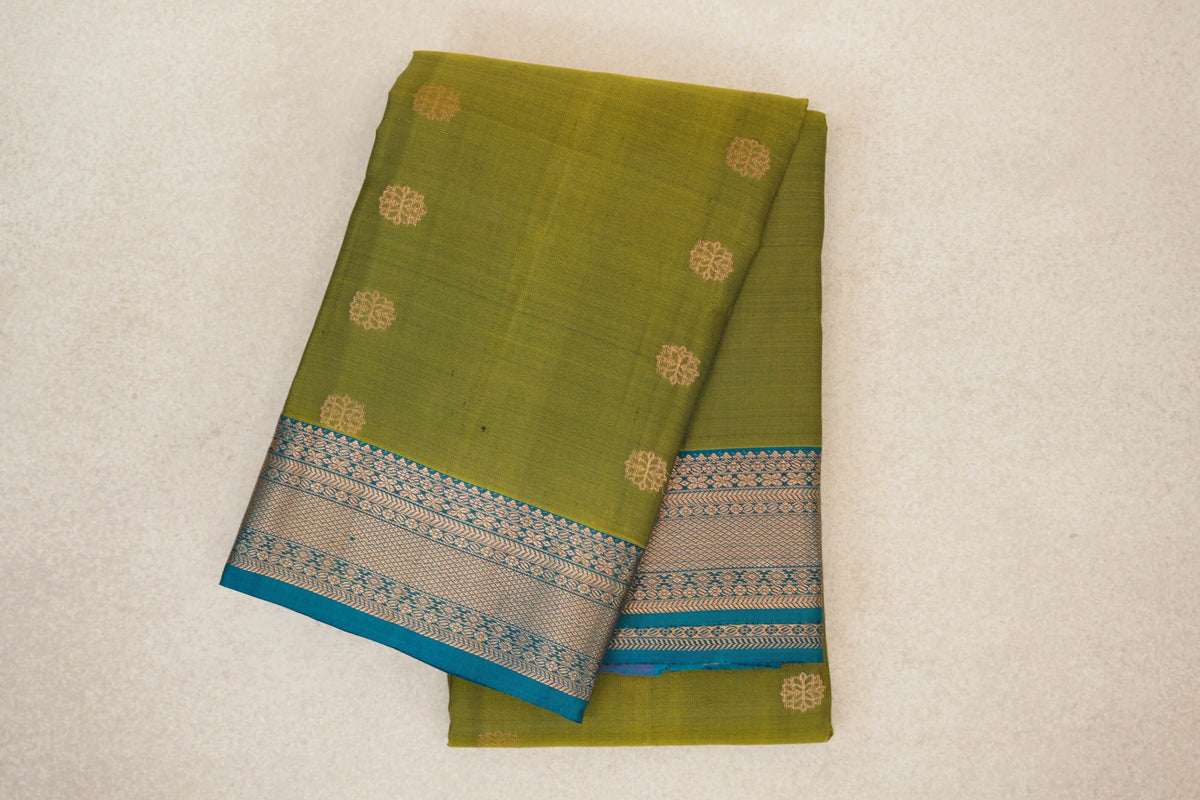 Folded olive green Kanchipuram silk saree with small gold-toned round motifs and a teal blue border woven with intricate patterns.