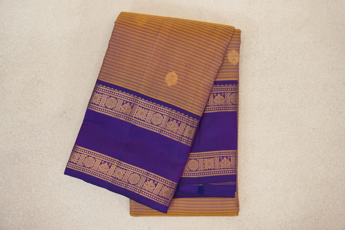 Folded mustard yellow Kanchipuram silk saree with fine purple stripes, scattered gold motifs and a deep purple border with floral bands.