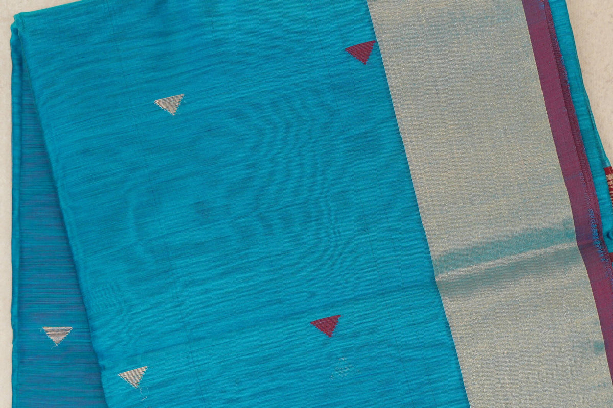 Folded Maheshwari silk cotton saree showing a bright turquoise blue body with small silver and deep red triangles beside a broad silver-toned border.