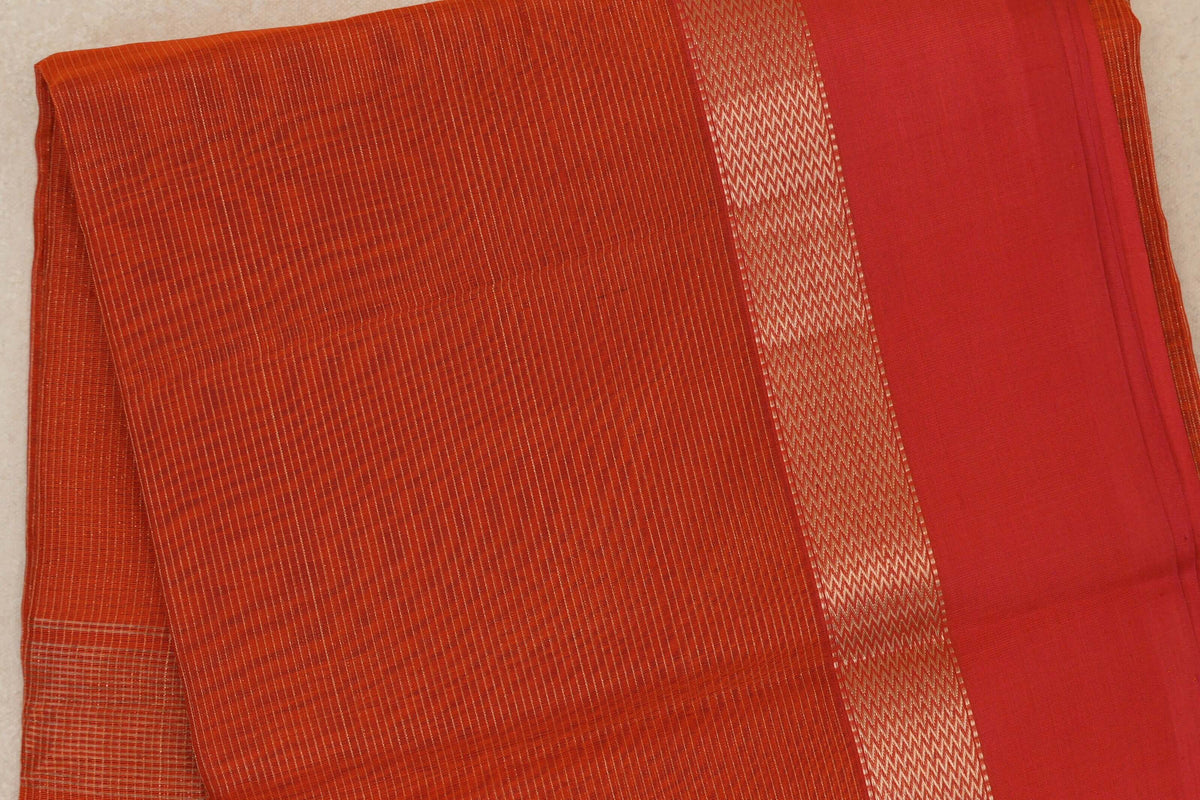 Folded Maheshwari silk cotton saree showing a rust orange body with fine woven lines and a wide red border edged in gold zigzags.