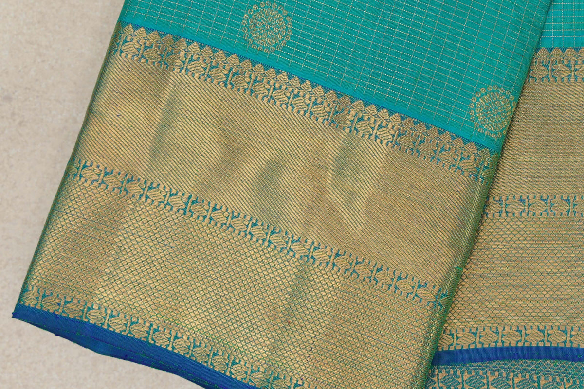 Close-up of a folded aqua green Kanjivaram silk saree, showing gold motifs and a wide zari border with fine geometric patterns and blue edging.