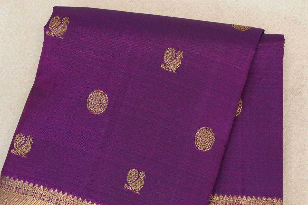 Folded purple Kanjivaram silk saree with gold peacock and circular motifs and a patterned gold border along the lower edge.