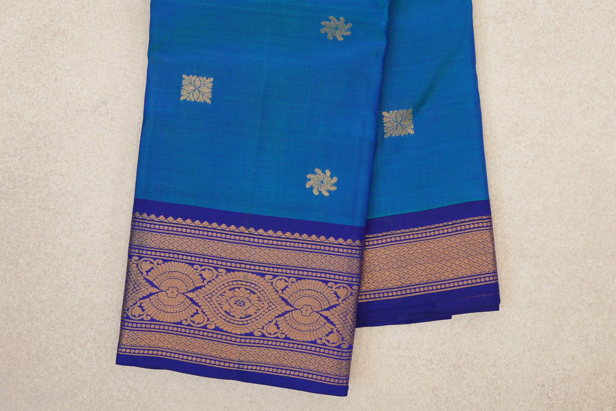 Folded light weight Kanchipuram silk saree with a peacock blue body, scattered gold-toned motifs and an ornate deep royal blue border.