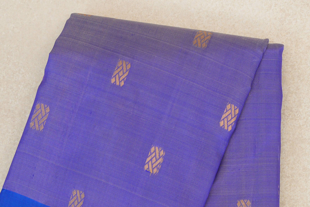 Folded lavender Kanchipuram silk saree with scattered gold-toned woven motifs and a royal blue border visible at the lower edge.