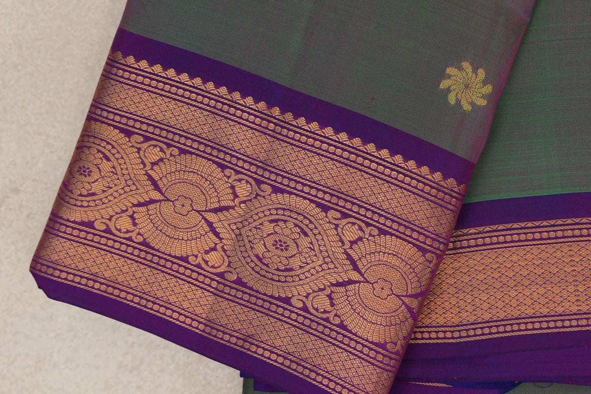 Folded light weight Kanchipuram silk saree showing a green body with a small gold motif and a wide violet purple border with gold floral and lattice patterns.