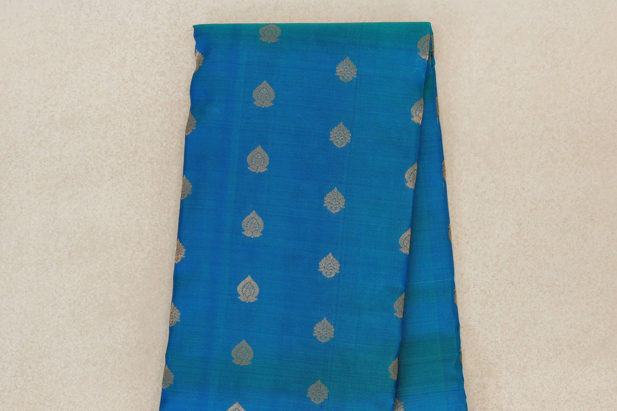 Folded peacock blue Kanchipuram silk saree showing the body’s scattered gold-toned leaf motifs and a layered edge.
