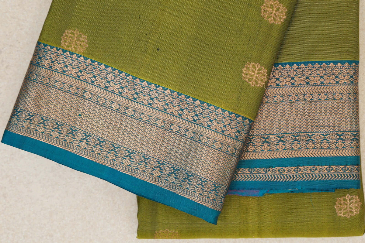 Folded olive green lightweight Kanchipuram silk saree with small gold-toned motifs and a wide teal blue border patterned with flowers and geometric designs.